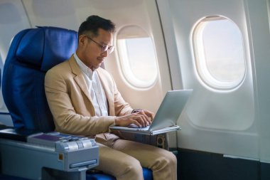 Şık takım elbiseli gülümseyen işadamı uçuş sırasında uçak kabininde dizüstü bilgisayarla çalışıyor..