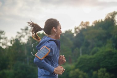 Gün batımında parkta koşarken müzik dinleyen kadın koşucunun yan görüntüsü.