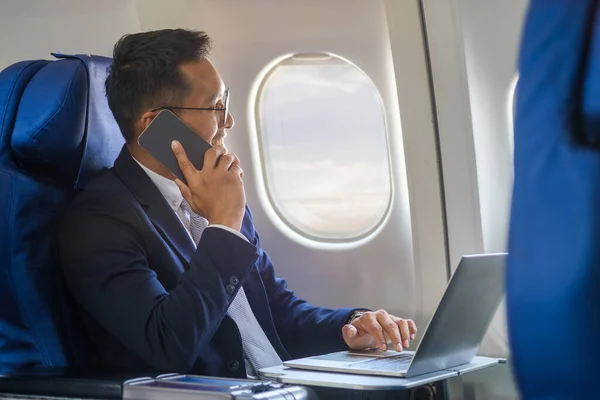Millennial businessman talking on mobile phone while sitting in airplane cabin with sun shining trough airplane window.