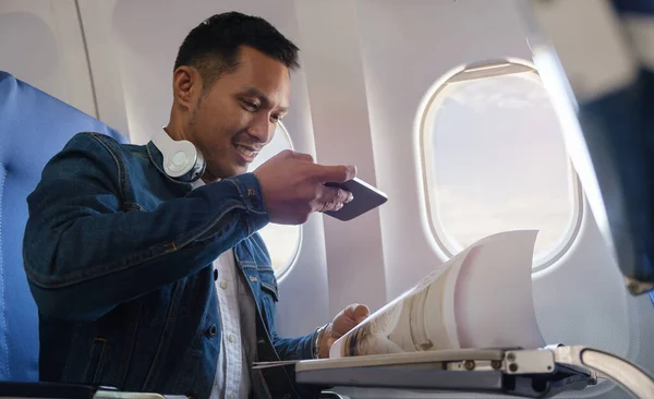 Smiling man in jean jacket using smart phone while sitting in airplane during flight. Traveling and technology