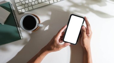Man hands holding smart phone on white office desk. Top view, blank screen for your advertise design.