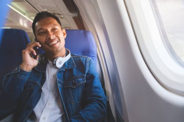 Smiling young asian man sitting next to window comfortable seat in aircraft cabin  during flight.