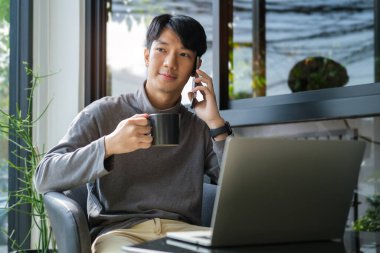 Young man wearing casual clothes drinking hot coffee and talking on a mobile phone at outdoor cafe.