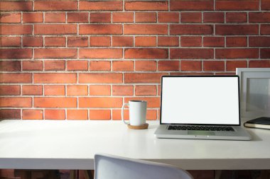 Front view of laptop, books, cup of coffee and stationery on white table against brick wall. Copy space for your text.