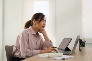 Thoughtful Asian woman employee looking away while using laptop at workstation.