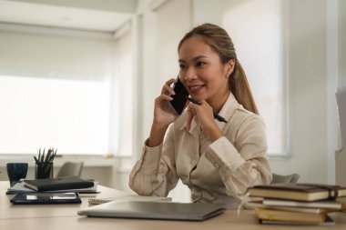 Confident female manager talking on a mobile phone while sitting at office desk with laptop computer.
