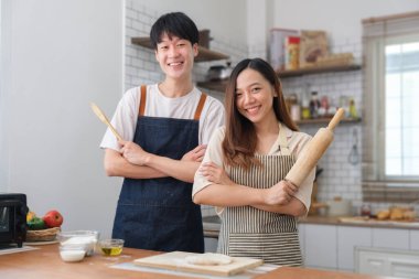 Image of lovely young couple in apron holding  holding rolling pins while in kitchen interior.