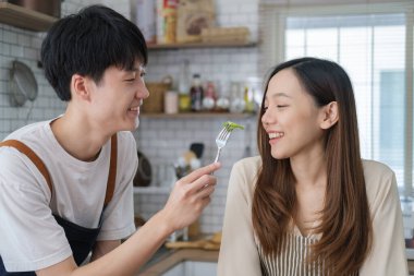 Romantic asian man feeding his girlfriend while cooking in kitchen, spending free weekend time together at home.