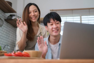 Happy young married couple  making video call via laptop while cooking in modern kitchen at home