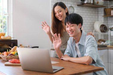 Friendly young married couple making video call on laptop sitting in modern kitchen at home.