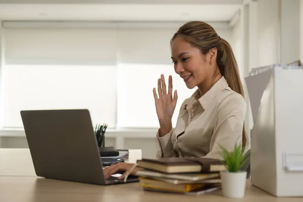 Attractive young businesswoman waving hand, having video call via laptop in her office.