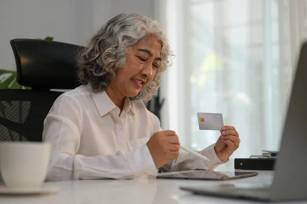 Happy retired woman holding credit  card doing online shopping, order delivery, booking tour on laptop.