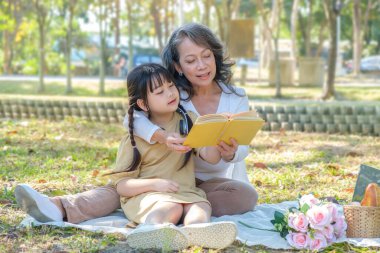 Caring senior woman embracing and reading fairytale to little grandchild during picnic in the park.