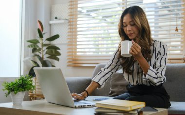 Çekici genç bir kadın sıcak kahve içiyor ve sıcak oturma odasında dizüstü bilgisayar kullanıyor..