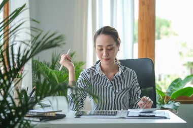 Beautiful young businesswoman working with digital tablet and analysing financial document at her office desk.	