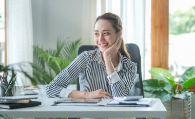 Satisfied asian young woman relaxing at her workplace and looking through the window at office.	