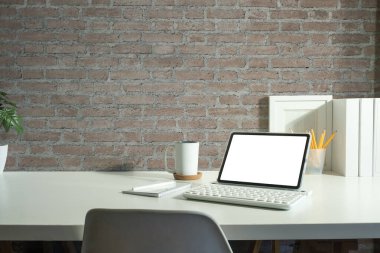 Beyaz masa üzerinde dijital tablet, resim çerçevesi, kitaplar ve ev bitkisi bulunan rahat işyerinin içi.