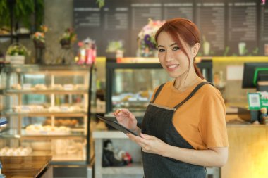 Kahve dükkanında dikilip dijital tablet kullanan önlük giymiş gülümseyen kadın garsonun portresi.