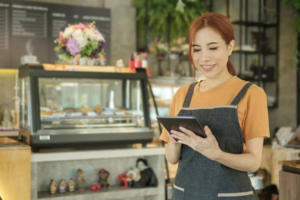 Smiling caucusian woman entrepreneur using digital tablet while standing in front of the counter in cozy coffee shop interior.