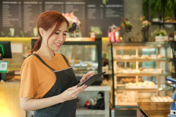 Smiling cafe owner standing in front of showcase and reading online order on digital tablet.