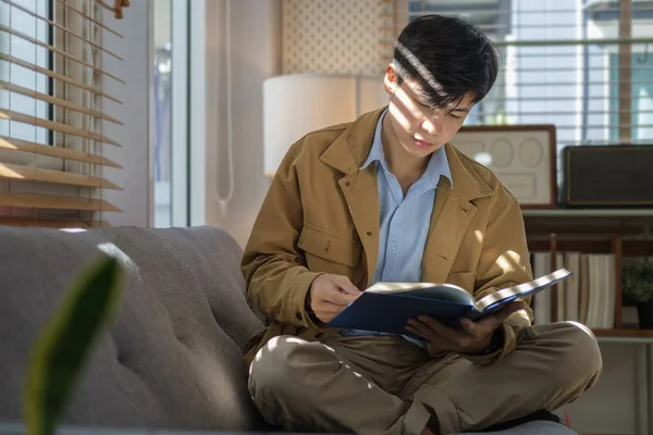 Positive Asian Male Reading Book While Sitting Couch Sunlight Blinds — Stock Photo, Image