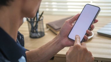 İş yerinde otururken akıllı telefonları elinde tutan genç adamın omuz üstü görüntüsü.