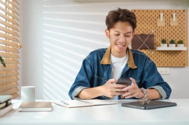 Hoş, genç Asyalı adam oturma odasında oturuyor ve cep telefonuyla sohbet ediyor..