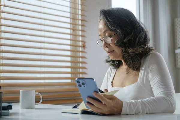 Starsza Kobieta Okularach Używająca Telefonu Komórkowego Siedząc Przytulnym Domu Laptopem — Zdjęcie stockowe