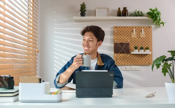 Relaxed asian man holding coffee cup and looking away while sitting in modern home office.