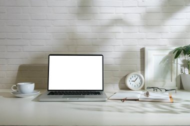 Boş ekran, resim çerçevesi, kahve fincanı ve beyaz masadaki not defteri olan dizüstü bilgisayarın ön görüntüsü.
