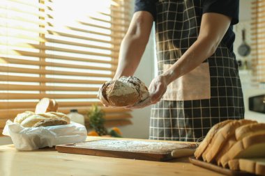 Erkek fırıncı elleri önlük takıyor ve elinde kaynak ekmeği var. Parlak mutfakta duruyor..