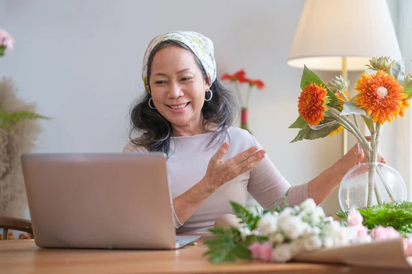 Smiling senior woman with bouquet of fresh flowers talking via video call on laptop. Elderly lifestyle and technology concept.