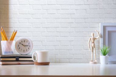 Bir fincan kahve, saat, resim çerçevesi ve kalem tutacağı tuğla duvara karşı beyaz masada..