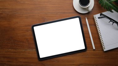 Beyaz ekranlı dijital tablet, kahve fincanı, bardaklar ve ahşap ofis masasında not defteri..