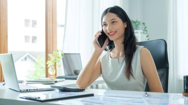 Neşeli Asyalı kadın dizüstü bilgisayarının önünde oturuyor ve cep telefonuyla konuşuyor..