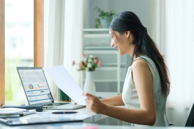 Genç Asyalı iş kadını işyerinde oturuyor ve mali belgeleri okuyor..
