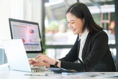 Genç Asyalı kadın ofis çalışanı parlak ofiste oturuyor ve bilgisayar bilgisayarı üzerinde çalışıyor..