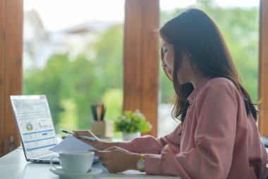 Genç Asyalı kadın ofis çalışanı parlak ofiste oturuyor ve bilgisayar bilgisayarı üzerinde çalışıyor..
