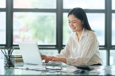 Young asian female office worker sitting in bright office and working on computer laptop.	