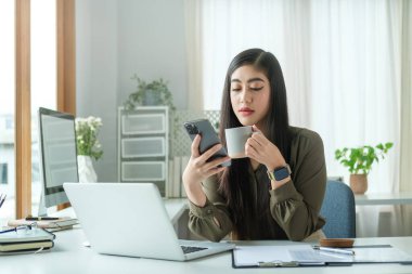 Kahve fincanı tutan ve modern ofiste cep telefonu kullanan çekici bir iş kadını..