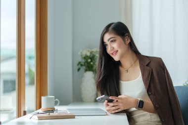 Tatmin olmuş Asyalı genç bir kadın iş yerinde dinleniyor ve ofisin penceresinden bakıyor..