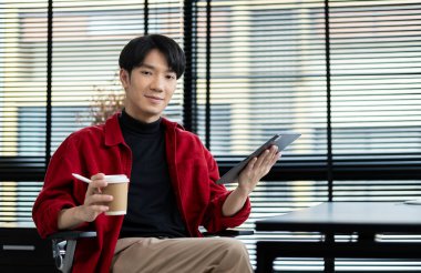 Girişimci iş adamı elinde bir fincan kağıt kahve tutuyor ve modern ofiste dijital tablet kullanıyor..