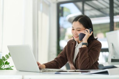 Neşeli Asyalı kadın dizüstü bilgisayarının önünde oturuyor ve cep telefonuyla konuşuyor..