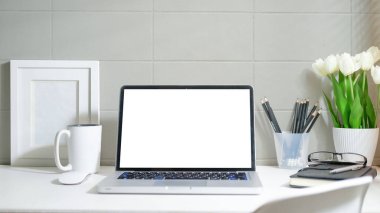 Laptop computer with empty screen, coffee cup, picture frame and stationery on white table.