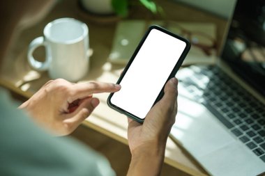 İş yerinde otururken akıllı telefonları elinde tutan genç adamın omuz üstü görüntüsü.