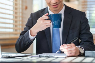 Gülümseyen genç işadamı kahve içiyor ve dijital tablet kullanıyor, masasında finansal ve pazarlama projeleri üzerinde çalışıyor.