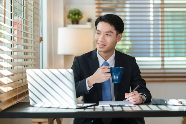 Relaxed asian man holding coffee and looking away while sitting in modern office.