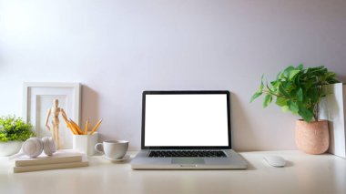 Boş ekranlı dizüstü bilgisayar, saksı bitkisi, resim çerçevesi ve beyaz masadaki kırtasiye malzemesi.