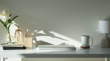 Laptop, kahve fincanı, lamba ve çiçek beyaz masada..