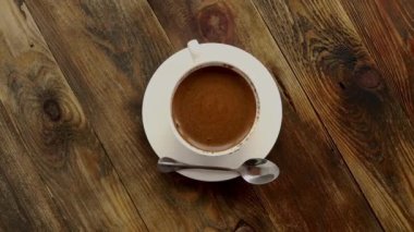 Çay kaşığı koyu ahşap masada dönen çay kaşığıyla çay tabağının üzerinde sade, beyaz bir fincan kahve. Üst görüntü, kopyalama alanı, yavaş çekim. Kapat..